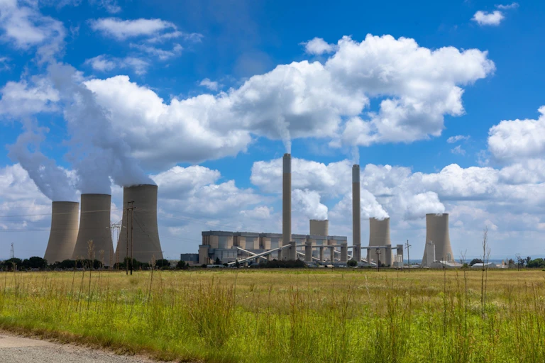 A coal fired power station in South Africa emitting smoky gas into the sky. There's a grass field in the foreground.