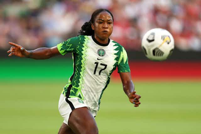 A woman in a green and white jersey puffs her cheeks with exertion as she pursues a soccer ball.