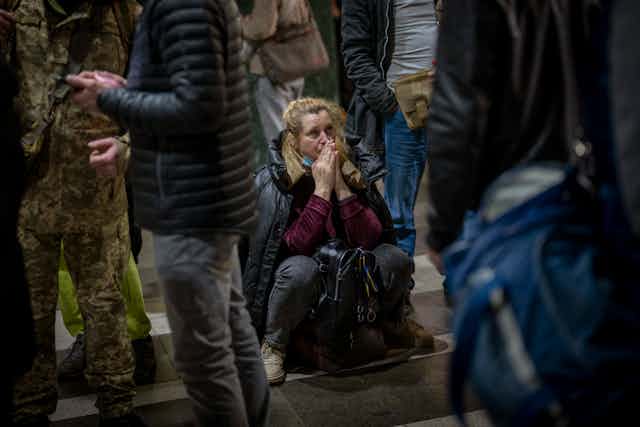 A woman sitting on the ground among a group of standing people holds her hands to her face as she waits for a train trying to leave Kyiv, Ukraine,