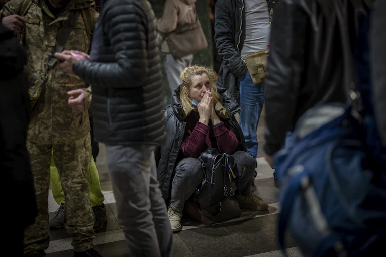 A woman sitting on the ground among a group of standing people holds her hands to her face as she waits for a train trying to leave Kyiv, Ukraine,