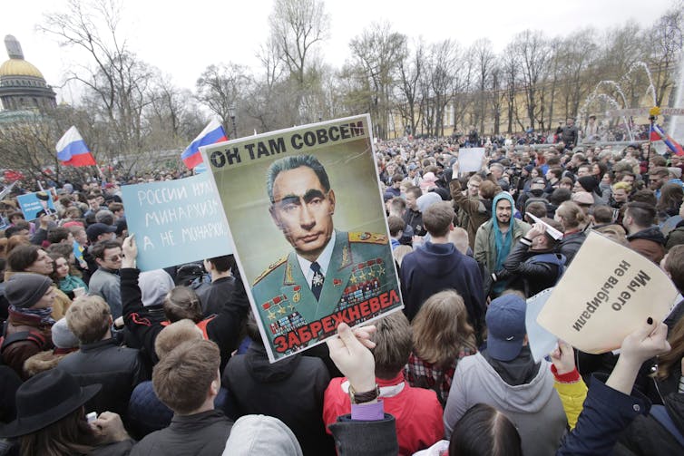 A crowd of protesters, one waving a sign of a dark haired man with thick eyebrows.