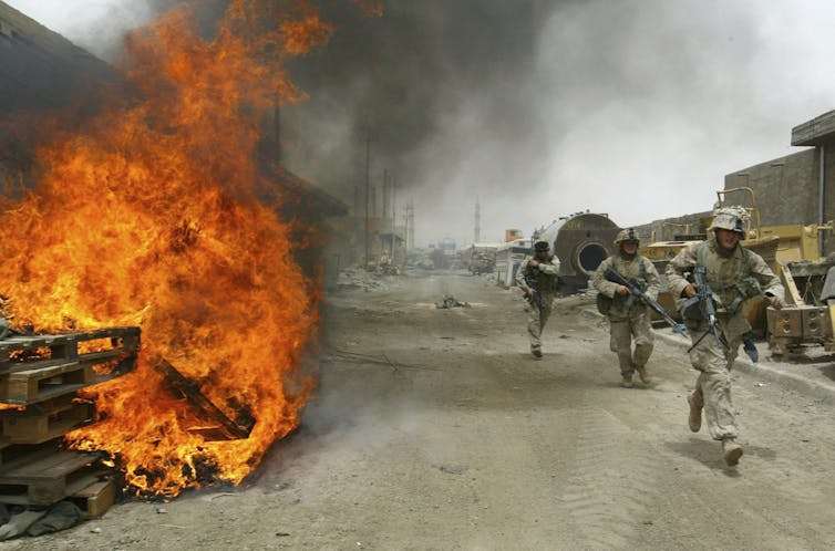 Large flames are seen as a line of armed soldiers walk by.