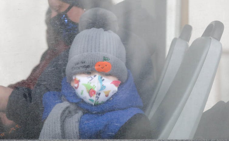 A child wearing woolly and face mask looks out of bus window.