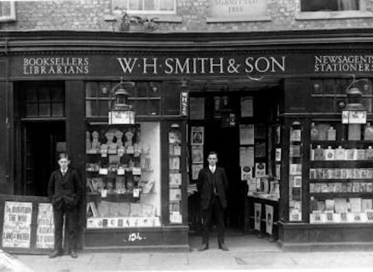 Old black and white photo of two men stood outside a shop.