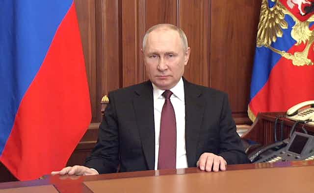Vladimir PUtin sits at a desk with the Russian flags behind him.