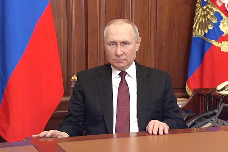 Vladimir PUtin sits at a desk with the Russian flags behind him.