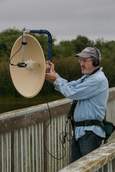 Man on bridge, wearing headphones, holding large dish-shaped microphone.