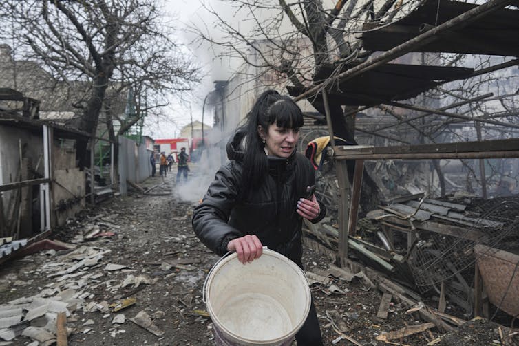 A black-haired woman holding a bucket walks past debris.