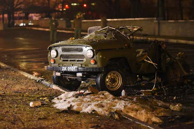 a small olive drab truck, it's front end intact, the rest mangled, in an otherwise empty parking lot at night