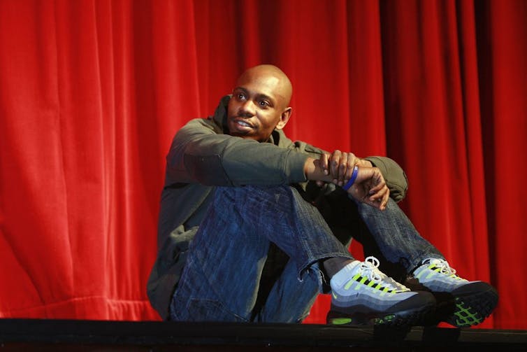 Man sitting on stage in front of red curtain.