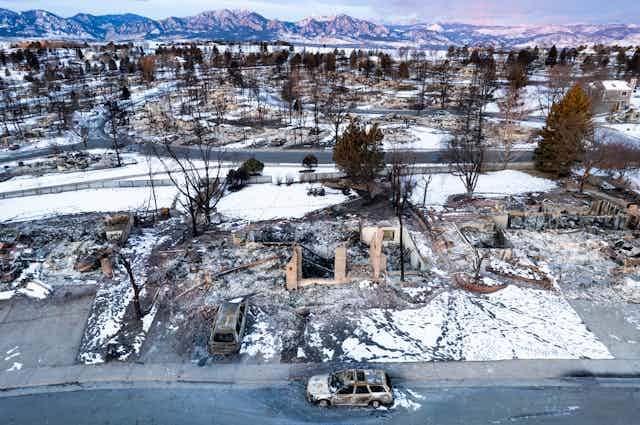A burned neighborhood in ruins with mountains in the background. House foundations, a few brick walls and and shells of burned-out vehicles remain.