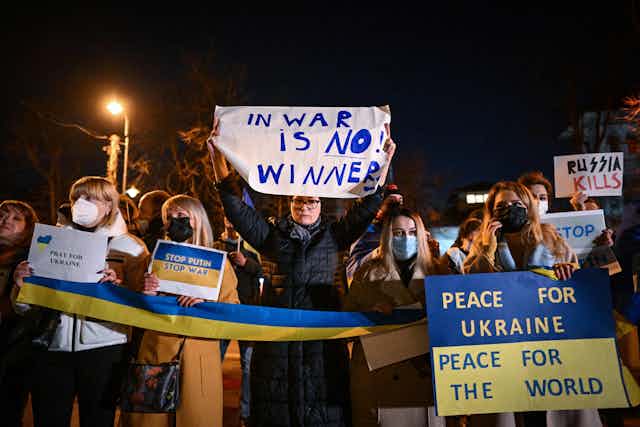 A few people stand together at nighttime and hold a long Ukrainian flag and a sign that says "In war there is no winner" and "Peace for Ukraine, peace for the world" 