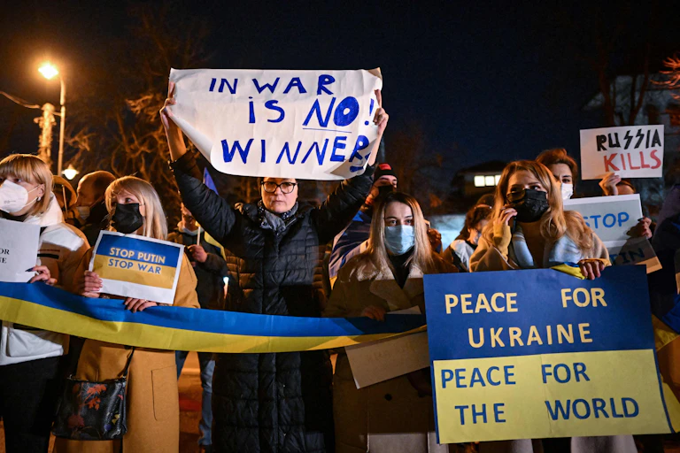 A few people stand together at nighttime and hold a long Ukrainian flag and a sign that says "In war there is no winner" and "Peace for Ukraine, peace for the world"