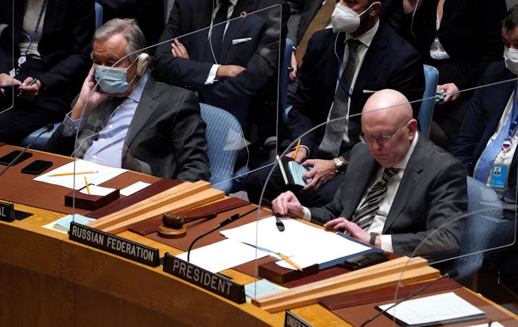 Dois homens, Antonio Guterres e o embaixador russo Vasily Nebenzya, estão sentados um ao lado do outro, com divisórias de plástico, ao redor de uma mesa do Conselho de Segurança da ONU.