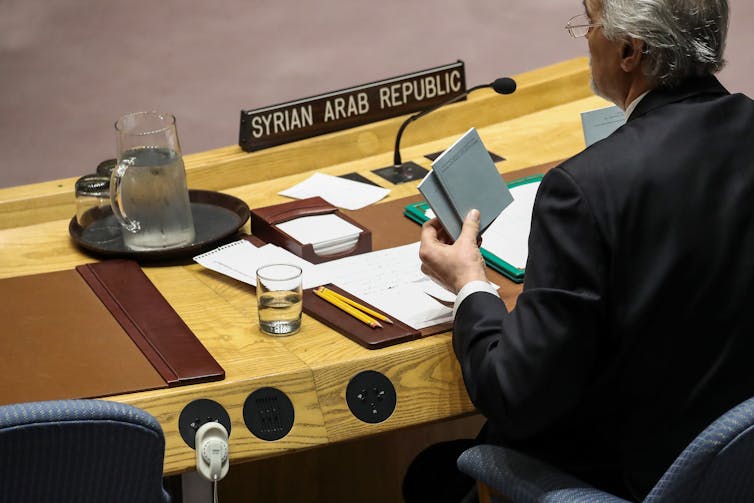 As costas de um homem são mostradas enquanto ele segura uma pequena Carta da ONU.  Ele está sentado em uma mesa da ONU com a placa da República Árabe Síria à sua frente.