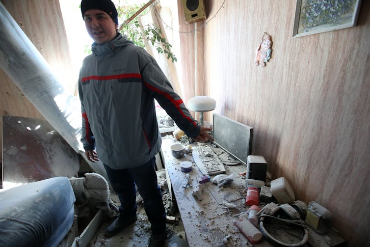 A man stands next to a table with a computer and keyboard, now destroyed with debris, in a heavily damaged looking home