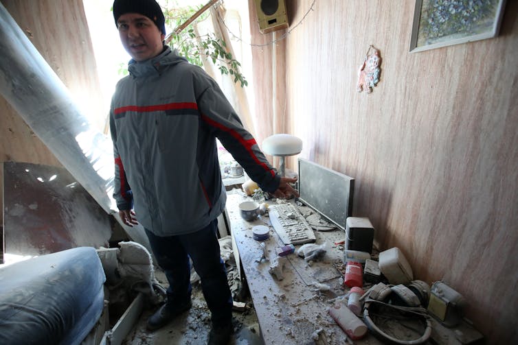 Um homem está ao lado de uma mesa com um computador e teclado, agora destruído com detritos, em uma casa com aparência muito danificada