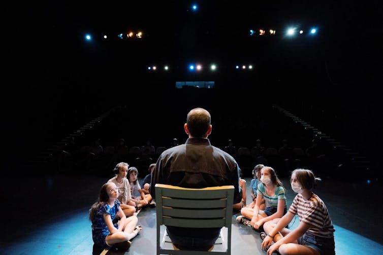 Children sit on the floor, listening to a man in a chair.