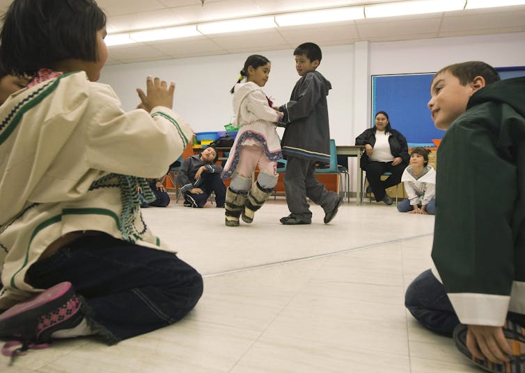 Two children watch and engage with one another as two other children dance in front of them.