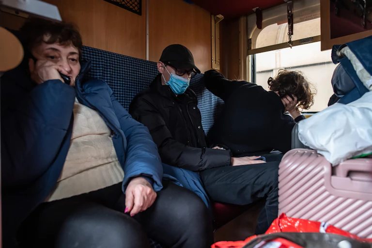 Three people with suitcases and bags in a dark train carriage. An older woman holds a phone to her ear while a young man wearing a mask rests and another person looks out the window.