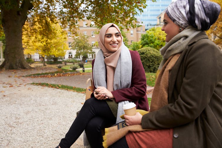 Two people sit on a park bench with takeaway coffee, smiling and talking.