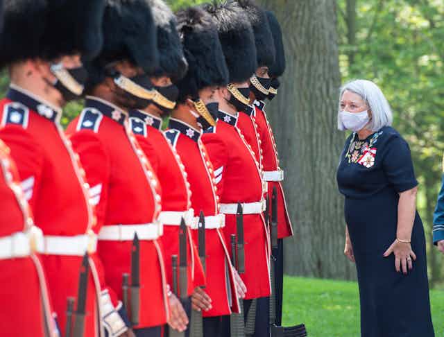 A woman with grey hair wearing a masks looks at a line of soldiers in bright red tunics and black fur hats.