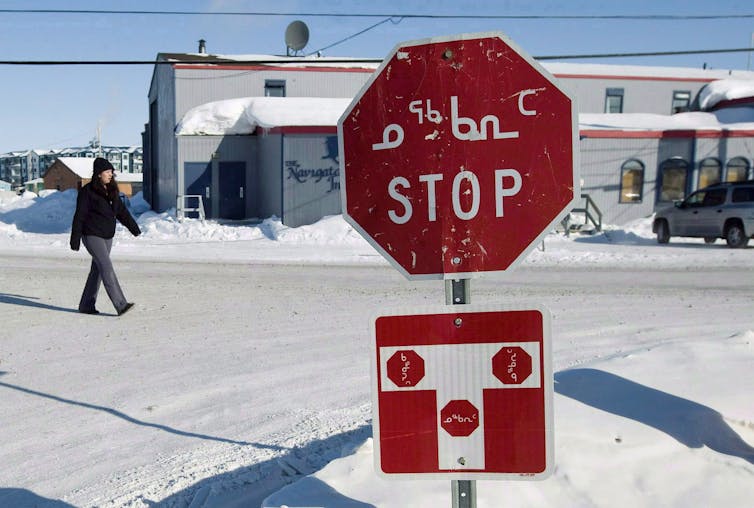 A woman in a hat and jacket walks past a stop sign displayed in both English and Inuktitut.