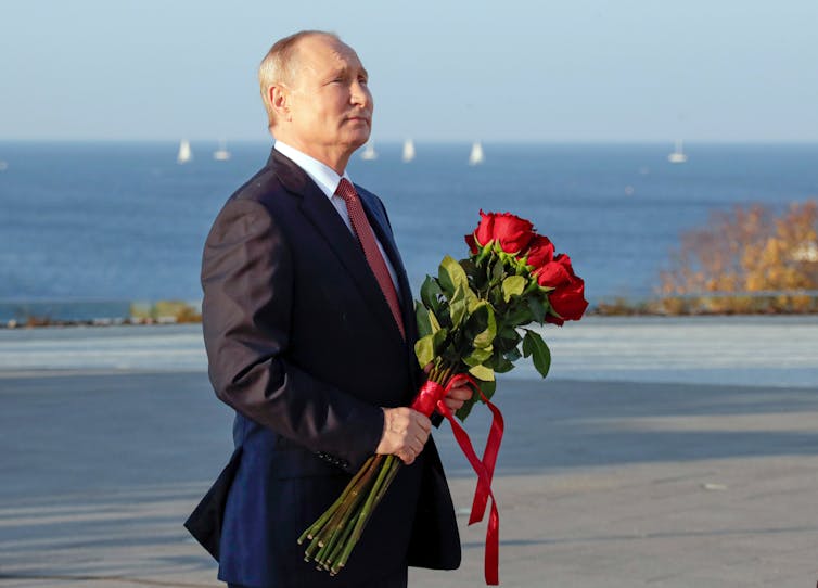 A bald man holds a bouquet of red roses with the sea and sailboats in the background.