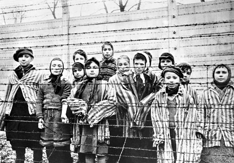 Children wearing striped prisoner clothing stand behind a barbed wire fence in this black-and-white photo