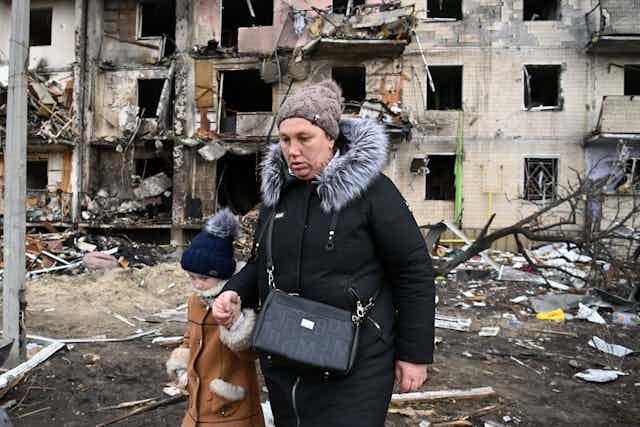 A woman walks with a child in front of a damaged residential building. 