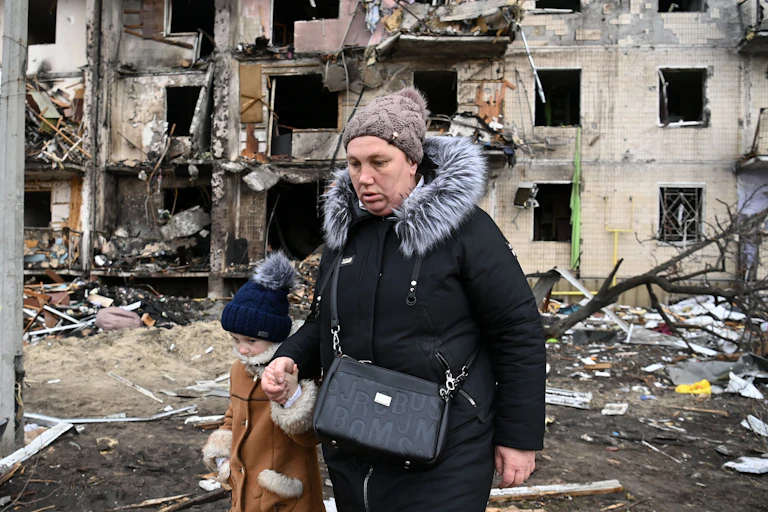 A woman walks with a child in front of a damaged residential building.