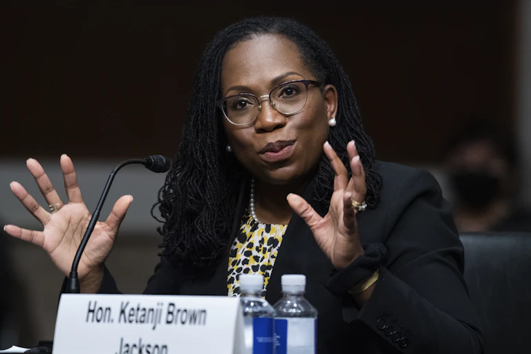 Ketanji Brown Jackson at a table with two bottles of water and gesturing with her hands as she speaks.