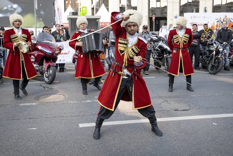 Men in bright red costumes and white bulbous hats carry swords and walk in formation on a street.