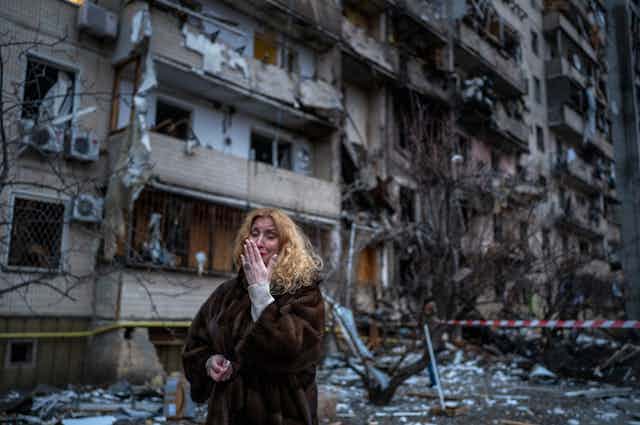 A woman puts her hand to her face as she walks in front of a destroyed building.
