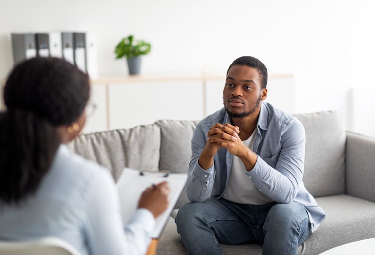 A man sitting on a sofa with his hands folded and a woman with a clipboard seen from behind