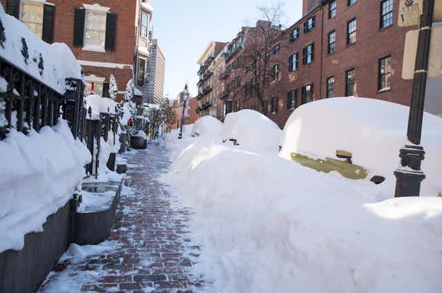 Snow covers cars and forms a tunnel where a sidewalk has been shoveled out