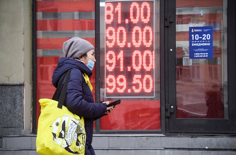 A woman carrying a yellow bag holds her phone as she walks down the street past a sign indicating how many rubles equals one dollar