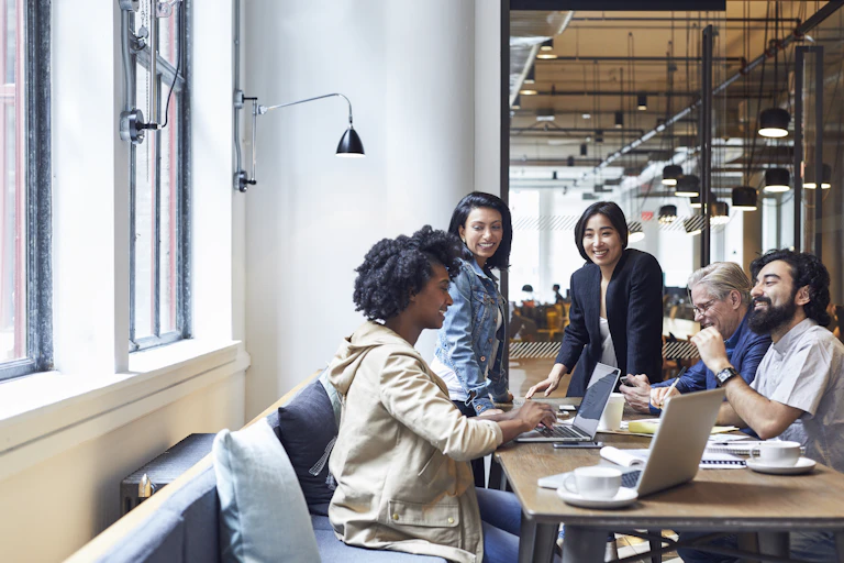 A diverse group of five people sit around a table smiling with laptops and papers in front of them
