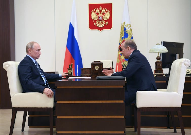 Two white men in dark suits face each other over a formal wooden desk.