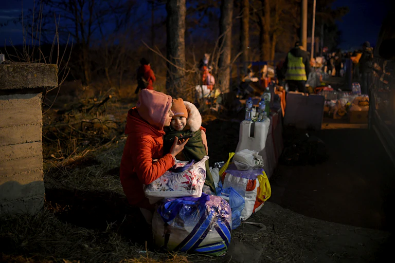A woman in winter clothing carrying a child and a large bag at night, with other people carrying bags in the background