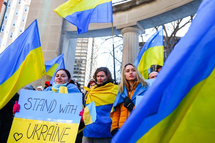 People waving Ukrainian flags and holding a sign painted like the Ukrainian flag reading'Stand with Ukraine'