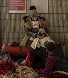 A woman holding a child on a bench against a tiled wall, with another child at her feet