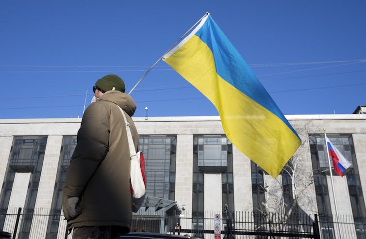 A person in winter clothing seen from behind, holding a Ukrainian flag outside a building with a Russian flag in front of it