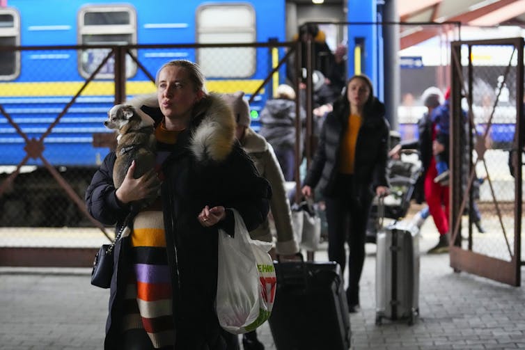 A woman in the foreground carrying a small dog and a suitcase in foreground, people from a blue train car behind her