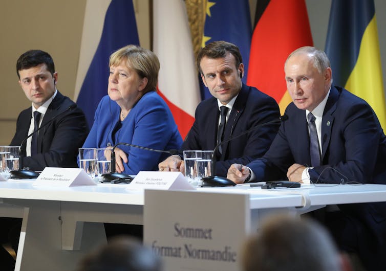 Volodymyr Zelensky, Angela Merkel, Emmanuel Macron and Vladimir Putin sit at a table with microphones