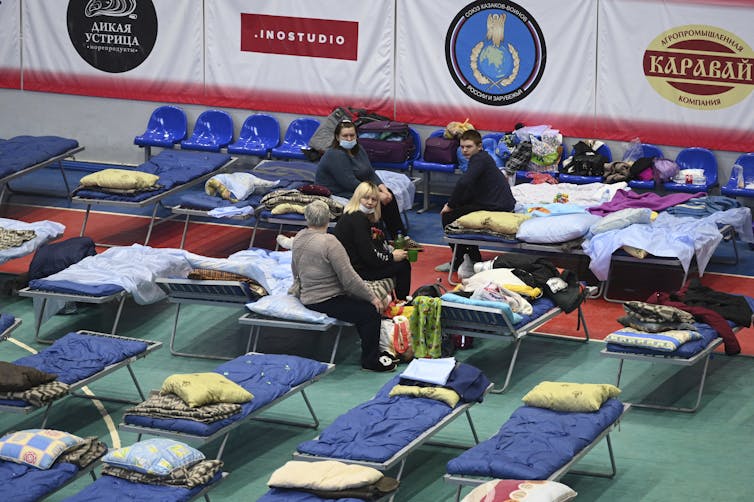 People sit on cots in a gymnasium.