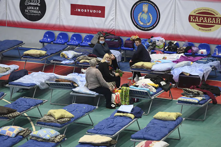 People sit on cots in a gymnasium.