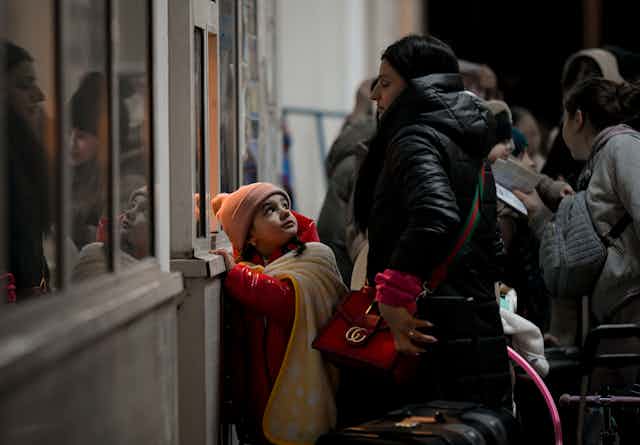 A little girl in an orange hat and red coat looks up at her mother as they stand in a line.