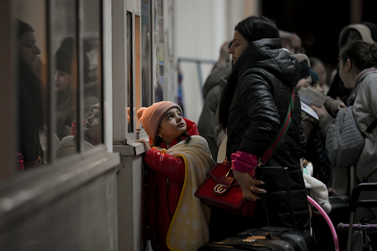 A little girl in an orange hat and red coat looks up at her mother as they stand in a line.
