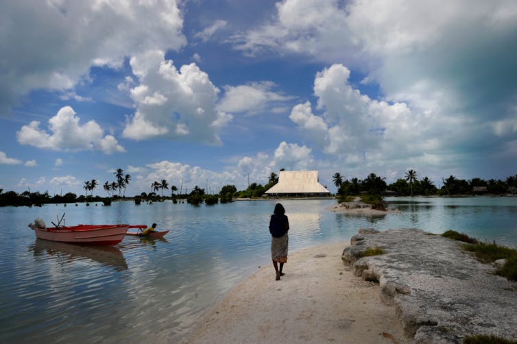 A woman stands at the edge of a flooded road looking at a home's roof across the water.